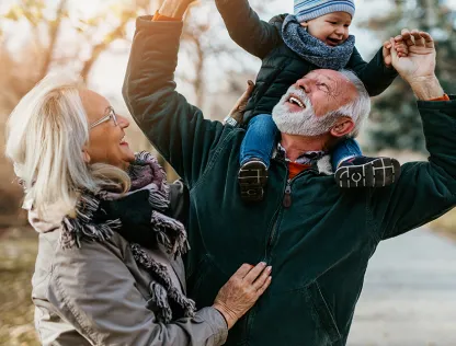 A man and a woman are smiling and looking up at their grandchild while the man carries the baby on his shoulders.