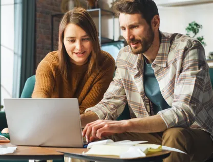 A man and a woman are looking at a laptop together, surrounded by a few papers.