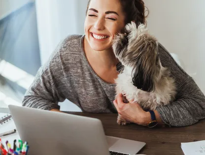 A woman is working on her laptop while her dog licks her face, and she smiles.