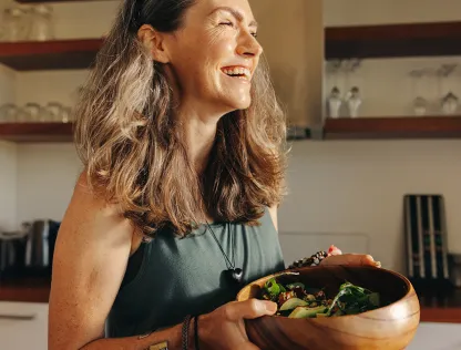 A woman is in the kitchen, holding a bowl with salad, and smiling.