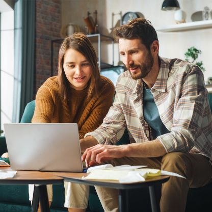 A man and a woman are looking at a laptop together, surrounded by a few papers.