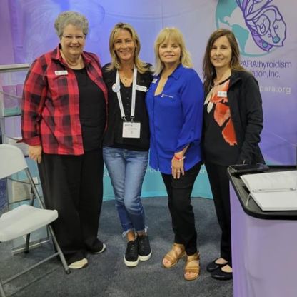 Four women are side-hugging each other and smiling at the camera at the office of the HypoPARAthyroidism Association Inc.