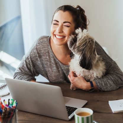 A woman is working on her laptop while her dog licks her face, and she smiles.