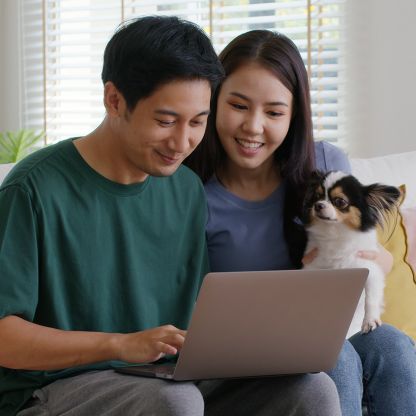 A woman and a man are looking at a laptop, smiling, while the woman holds a small dog.