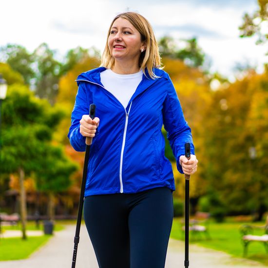 A woman is walking through a park with trekking poles while smiling.