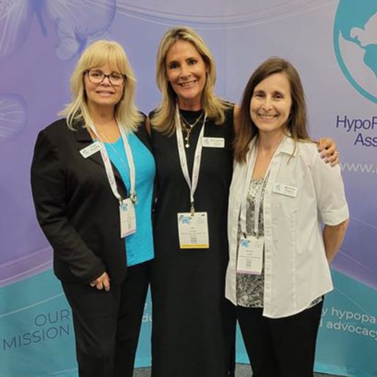 Three women from the HypoPARAthyroidism Association Inc. team are smiling and posing in front of the association's banner.