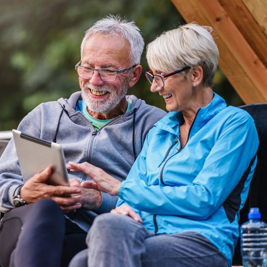 A man and a woman are sitting on a park bench, looking down at the tablet the man is holding, and smiling