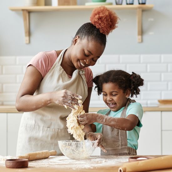 A woman and a toddler are making dough in the kitchen, with flour surrounding them, and both of them are smiling.