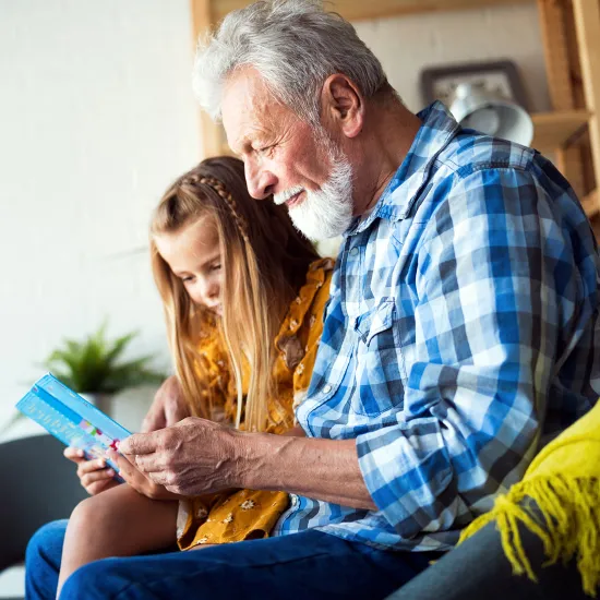A man and his granddaughter are sitting down and reading a children's book together.